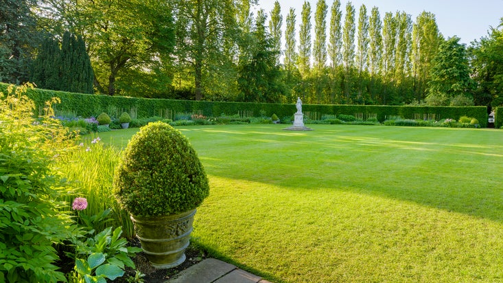 A photo showing a herbaceous border in early summer with flowers starting to bloom. There is a statue in the centre of a lawned area.